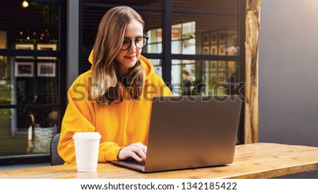 Front view. Girl in yellow hoodie sits in cafe at table in front of open laptop computer. Woman works on computer, checks email. Freelancer works remotely. Online education for adult. Lifestyle.