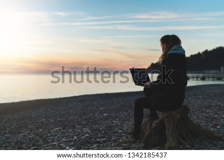 Back view. Woman in jacket sits on beach, working with laptop. Remote work. Work onlne from anywhere in world. Silhouette of girl with computer. Hipster girl working remotely. Lifestyle.