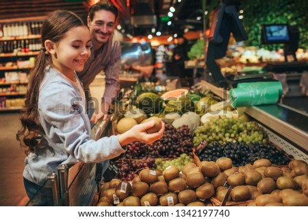 Young parent and daughter in grocery store. Father look at child and smile. Daughter hold kiwi in hands and smile. Positive happy picture.