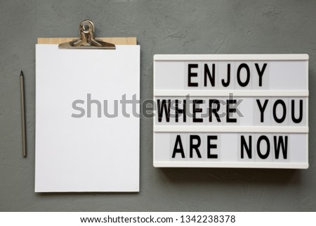 'Enjoy where you are now' words on modern board, clipboard with blank sheet of paper over concrete surface, top view. Flat lay, from above. 