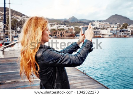Middle age Caucasian hipster female taking a landscape photo with her smartphone on the wooden harbor near the sea
