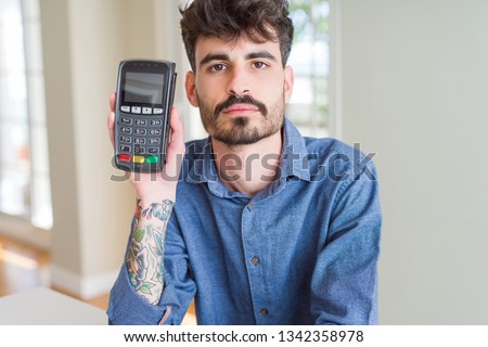 Young man holding dataphone point of sale as payment with a confident expression on smart face thinking serious