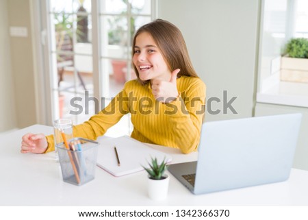 Beautiful young girl studying for school using computer laptop doing happy thumbs up gesture with hand. Approving expression looking at the camera showing success.