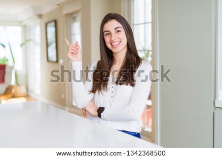 Beautiful young woman sitting on white table at home with a big smile on face, pointing with hand and finger to the side looking at the camera.
