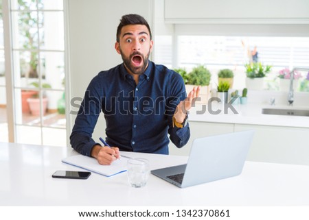 Handsome hispanic man working using computer and writing on a paper very happy and excited, winner expression celebrating victory screaming with big smile and raised hands