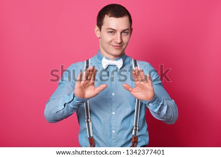 Young attractive male with short hairstyle. Model raises palms and shows stop sign, wears blue shirt with suspenders and white bow tie, isolated over pink concrete wall. Body language concept.