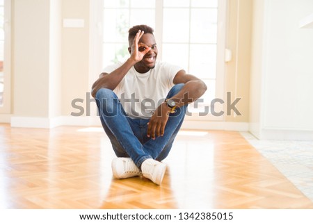 Handsome african american man sitting on the floor at home doing ok gesture with hand smiling, eye looking through fingers with happy face.