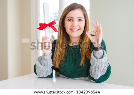 Beautiful young woman holding degree with red bow doing ok sign with fingers, excellent symbol