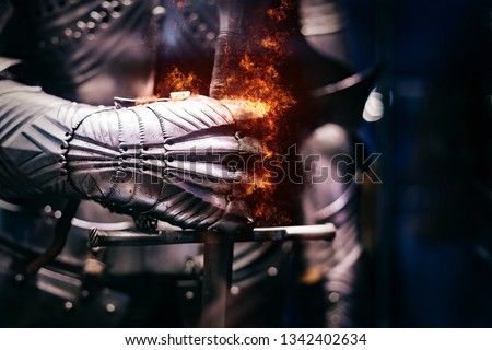 Close up of a Medieval steel armour with iron glove hand bursting with flames of fire, holding a giant sword Royalty-Free Stock Photo #1342402634