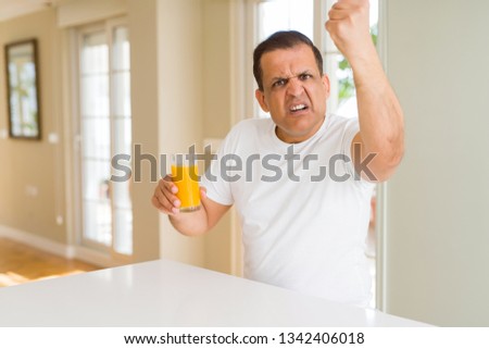 Middle age man drinking a glass of orange juice at home annoyed and frustrated shouting with anger, crazy and yelling with raised hand, anger concept