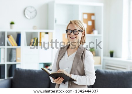 Young successful professional psychologist making notes in notepad while standing in front of camera inside office