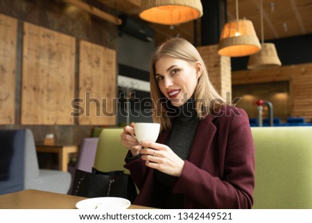 Beautiful smiling woman drinking coffee at cafe. Portrait of blonde  woman in a cafeteria drinking hot cappuccino and looking at camera. Pretty woman with cup of coffee.