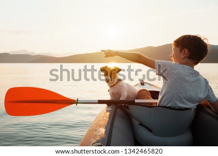 Happy boy with his dog Jack Russell Terrier paddling an inflatable kayak on the water mountain lake against the backdrop of beautiful orange sunset. Family sports vacation. Lens flare. Pet