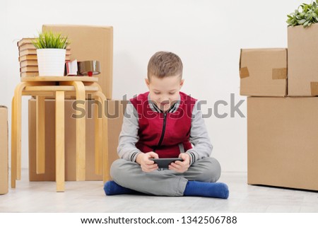 The boy child sits on the floor and plays on the smartphone. Boxes with cargo on a white background. The concept of moving to a new home.