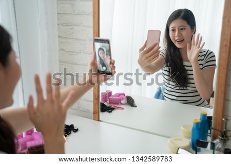 Beauty asian blogger filming makeup tutorial with smartphone in front of mirror at dressing table indoors. young woman waving hands starting recording video get ready in the morning bright bedroom.