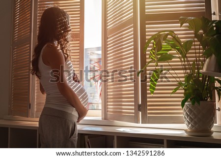 Beautiful modern pregnant woman with long curly hair, embracing a big tummy, standing by the window with shutters indoor and looking outside.