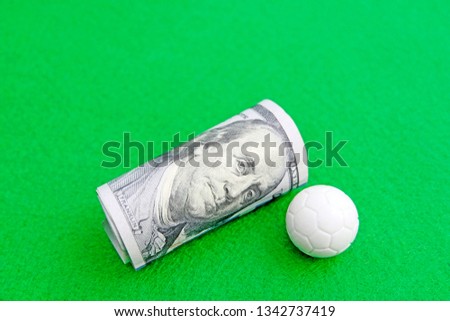 Football ball near the twisted hundred dollar bills of the United States. Green background. The concept of sports betting or competition for money, financial risk or corruption.