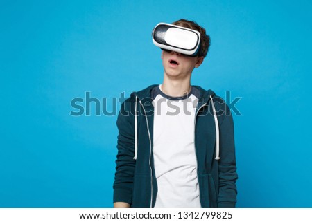 Portrait of amazed young man in casual clothes standing looking on headset isolated on blue wall background in studio. People sincere emotions, lifestyle concept. Mock up copy space. Advertising area