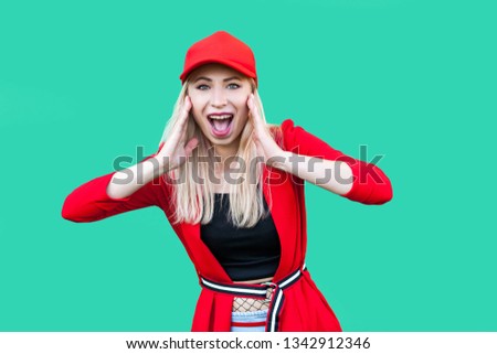 Portrait of amazed beautiful blond young hipster woman in red blouse and cap, standing, touching her face and looking at camera with suprised face. indoor studio shot, isolated on green background.