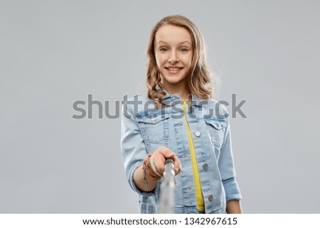 technology and people concept - happy smiling teenage girl taking picture by selfie stick and showing ok over grey background