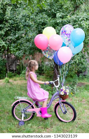 Little girl is riding on pink bicycle with balloons and wicker basket with bouquet of summer flowers. Kid gets gift. Celebration of Happy Birthday children party outside in summer garden in backyard.