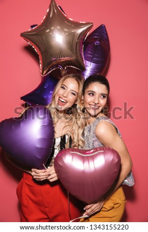 Image of two gorgeous girls 20s in stylish outfit laughing and holding festive balloons isolated over red background