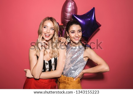 Image of two adorable girls 20s in stylish outfit laughing and holding festive balloons isolated over red background