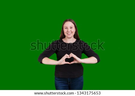 Young women standing gesturing a heart love sign with hands inviting smile on green screen background  