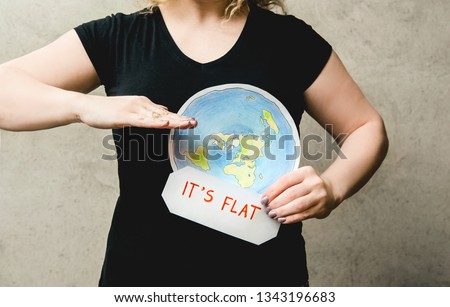 Flat Earther concept. Person who believes that Earth is flat disc. Anonymous woman holding flat Earth model in front of body with text: It`s flat. Isolated on gray background, studio shot. 