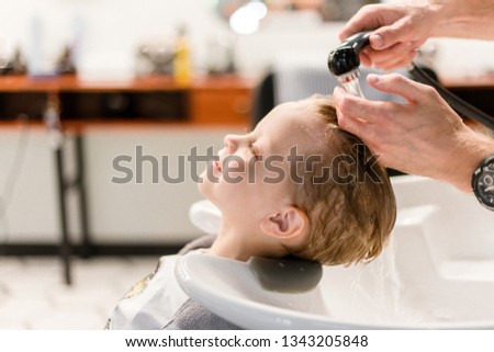 Hairdresser washing the child's hair under the water in the barbershop side view. Joyful child gets pleasure from washing hair