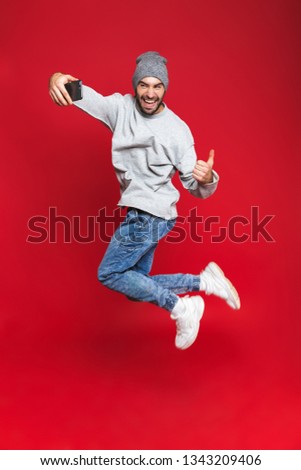 Full length photo of energetic man 30s in casual wear jumping and taking selfie photo on cell phone isolated over red background