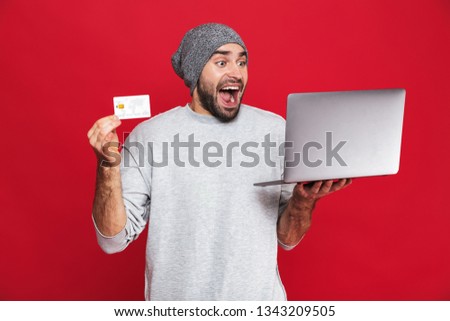 Photo of optimistic guy 30s in casual wear holding credit card and silver laptop isolated over red background