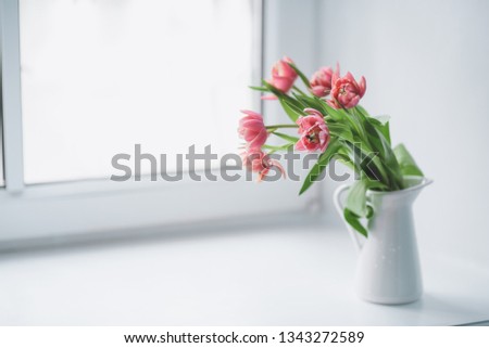 Pink peony tulips in a white jug stand on a window with a white window sill.