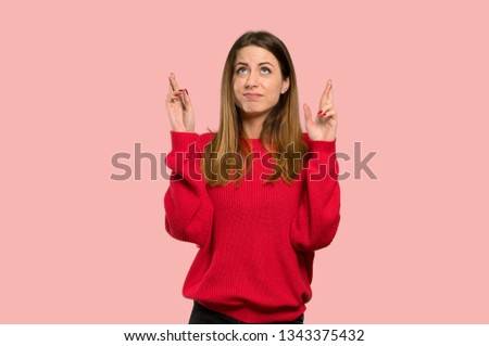 Young woman with red sweater with fingers crossing and wishing the best over isolated pink background
