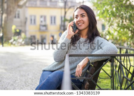 Smiling woman talking on phone and relaxing on bench outdoors. Pretty young lady sitting on bench with walkway and building in background. Urban lifestyle and communication concept. Front view.