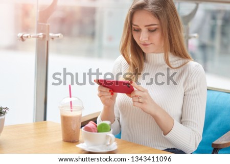 Image of young good looking blonde female sits in cafe, takes photo of her dinner with her phone, eats colourful macaroons and drinks milshake. Charming girl looks down, at screen. Technology concept.