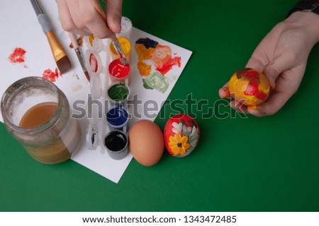 two hands drawing an egg, near them a paper on it gouache, two paint brushes and a water jar, near them two eggs on a green background