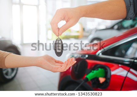 Young family buying first electric car in the showroom. Close-up of male hand giving car key to female hand on battery electric car background. Eco car sale concept Royalty-Free Stock Photo #1343530823