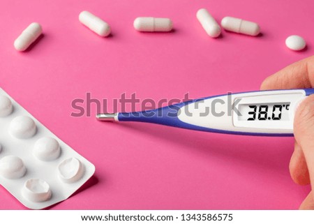 Electronic thermometer and pills on a pink background. High temperature 38 degrees Celsius on display. Copy space.