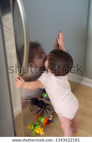 Gender neutral caucasian baby kissing reflection in stainless steel refrigerator door
