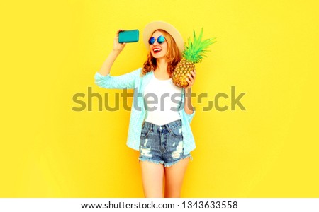 happy smiling woman with pineapple taking selfie picture by phone in summer round hat, sunglasses, shorts on colorful yellow background