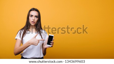 Beautiful young caucasian female with dark long hair looking at camera surprised while pointing at her smartphone dressed in white shirt against yellow background.