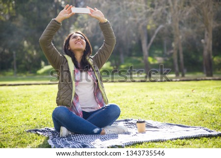 Delighted student girl taking selfie on phone outdoors. Young woman in casual jacket resting on plaid in park, and photographing sky on her smartphone. Picturing concept