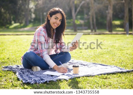 Cheerful female student working on class project. Beautiful girl in casual sitting on grass in park, holding tablet, taking notes and smiling at camera. Studies outdoors concept