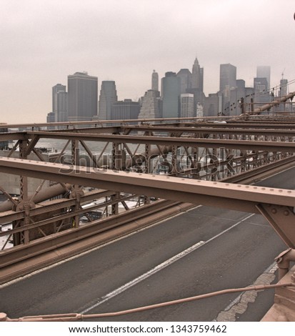 Beams and empty road on Brooklyn bridge with Manhattan skyline in background with high rise buildings and skyscrapers under cloudy grey sky and fog over cityscape