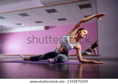 Fit young Caucasian brunette in sportswear sitting on mat in gym and doing side stretching. In background mirror.