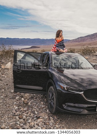 Brunette girl with long hair in a skirt sitting in the hatch of a black car in the middle of the road in the Valley of Death waving the American flag
