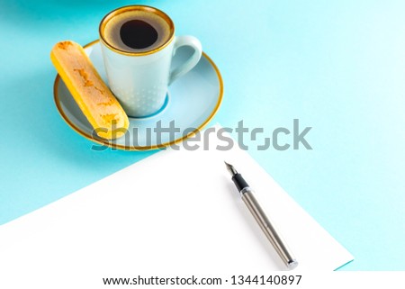 A sheet of white paper is empty on a blue background with an ink pen ,there is also a blue cup with coffee on a saucer and a plate with Savoyardi biscuits. Place for text.