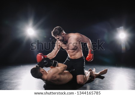 strong mma fighter in boxing gloves punching opponent while sportsman lying on floor