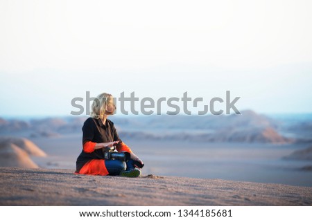 Caucasian white blond woman holding camera and sitting on a sand and looking to the desert in soft morning light. Photographer holding camera in her hands relaxing in the desert. Pretty woman  